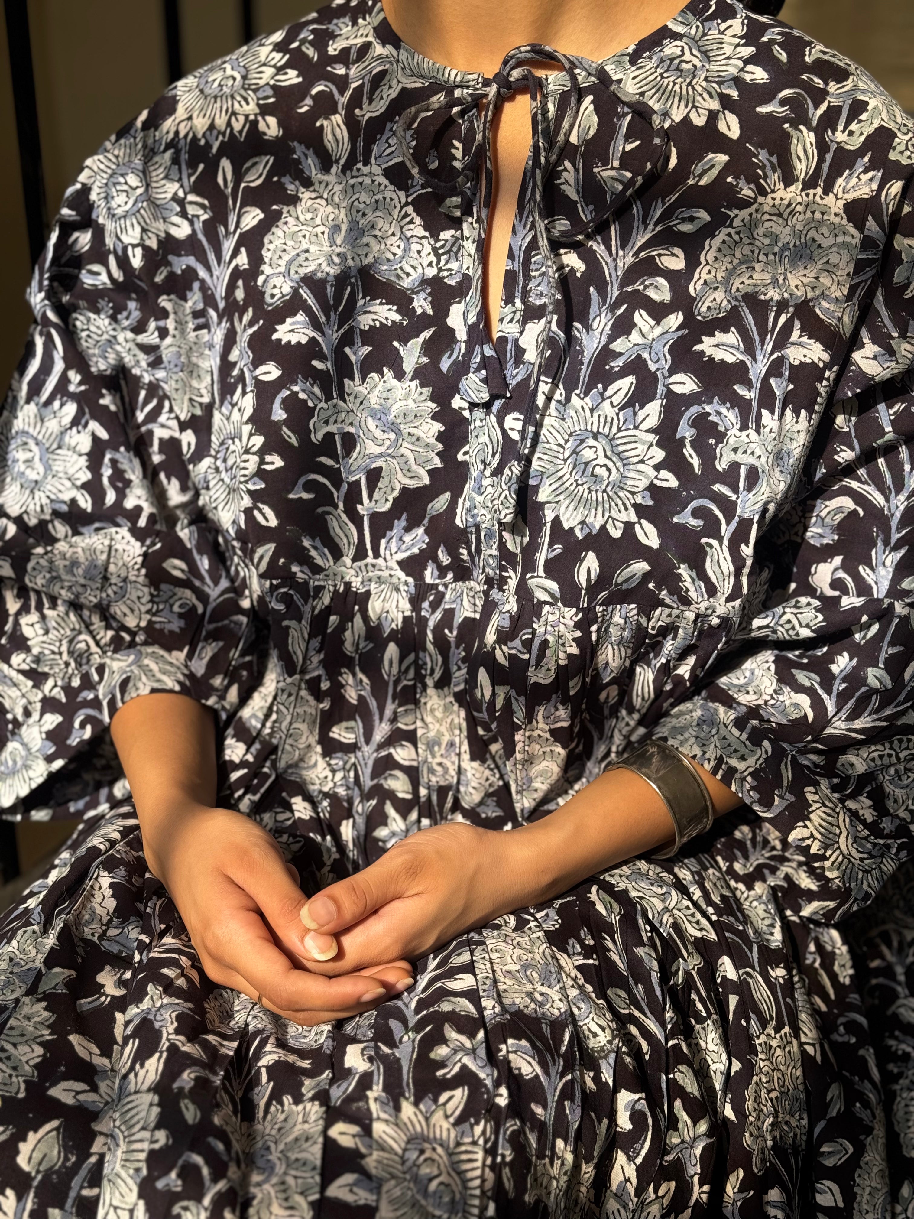 A close-up of a person wearing a loose, long-sleeve dress with a dark navy or black base and an intricate light blue floral and botanical print. The dress features a tie-neck detail and a gathered waist.